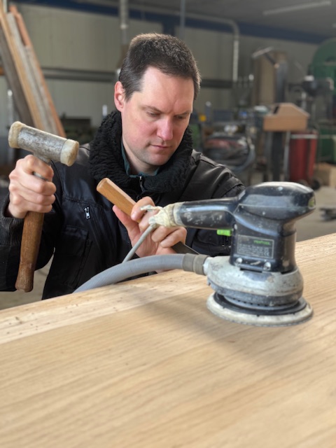 Bram die massieve eiken boomstamtafel schuurt en afwerkt in atelier